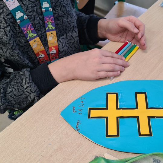 Ant suolo padėtas mergaitės pagamintas mėlynas herbas su geltonu Jogailos kryžiumi.