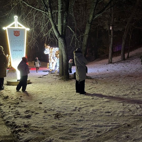 Kalnų parko snieguotas takas. Juo eina pora žmonių. Aplink šviečia kalėdinės šveiselės ir Lumina park instaliacijos.