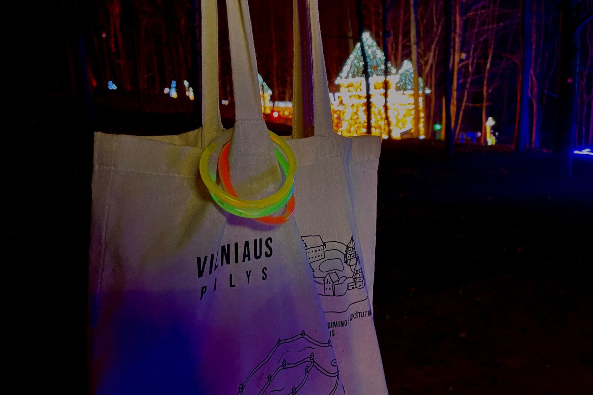 Vilniaus pilių medžiaginis krepšelis su žibančiomis trispalvės apyrankėmis. Fone matosi žiemiškas Lumina parkas.