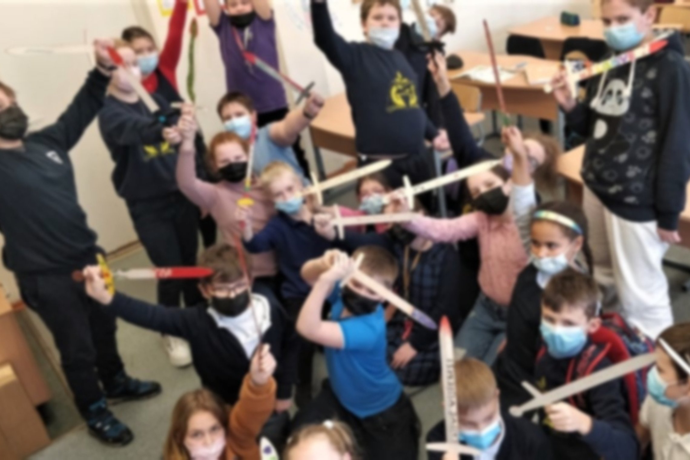 Mokiniai su mediniai kalavijais pozuoja aršiai nuotraukai klasėje.