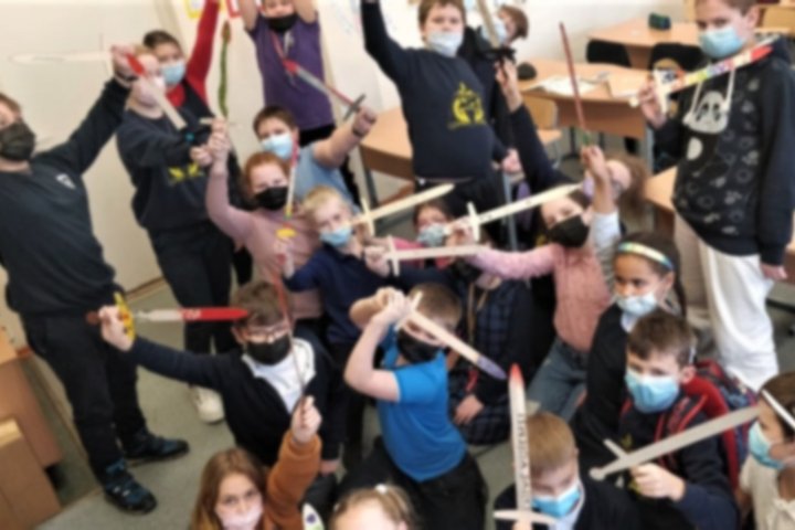 Mokiniai su mediniai kalavijais pozuoja aršiai nuotraukai klasėje.