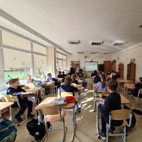Mokiniai nufotografuoti, sėdintys klasės suoluose, iš klasės galo.