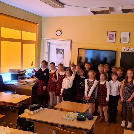 Mokinukai, sustoję klasės priekyje, mojuoja trispalvėm vėliavėlėmis.