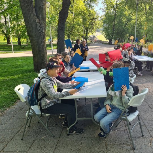 Mokiniai mojuoja spalvotais segtuvais, susėdę prie stalų Pilių parke.