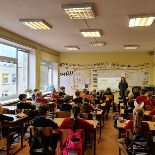 Klasė iš galo. Klasės priekyje edukatorė, dėstanti poamoką. Mokiniai žiūri į ją.