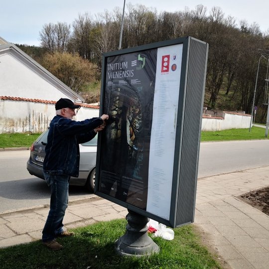 Darbuotojas Stanislovas montuoja naują "Initium Vilnensis" simpoziumo plakatą šveisdėžėje.