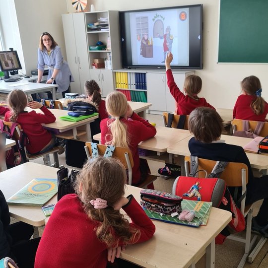 Kol kiti vaikai sėdi suoluose, viena mergaitė kelia ranką, nori užduoti klausimą edukatorei.