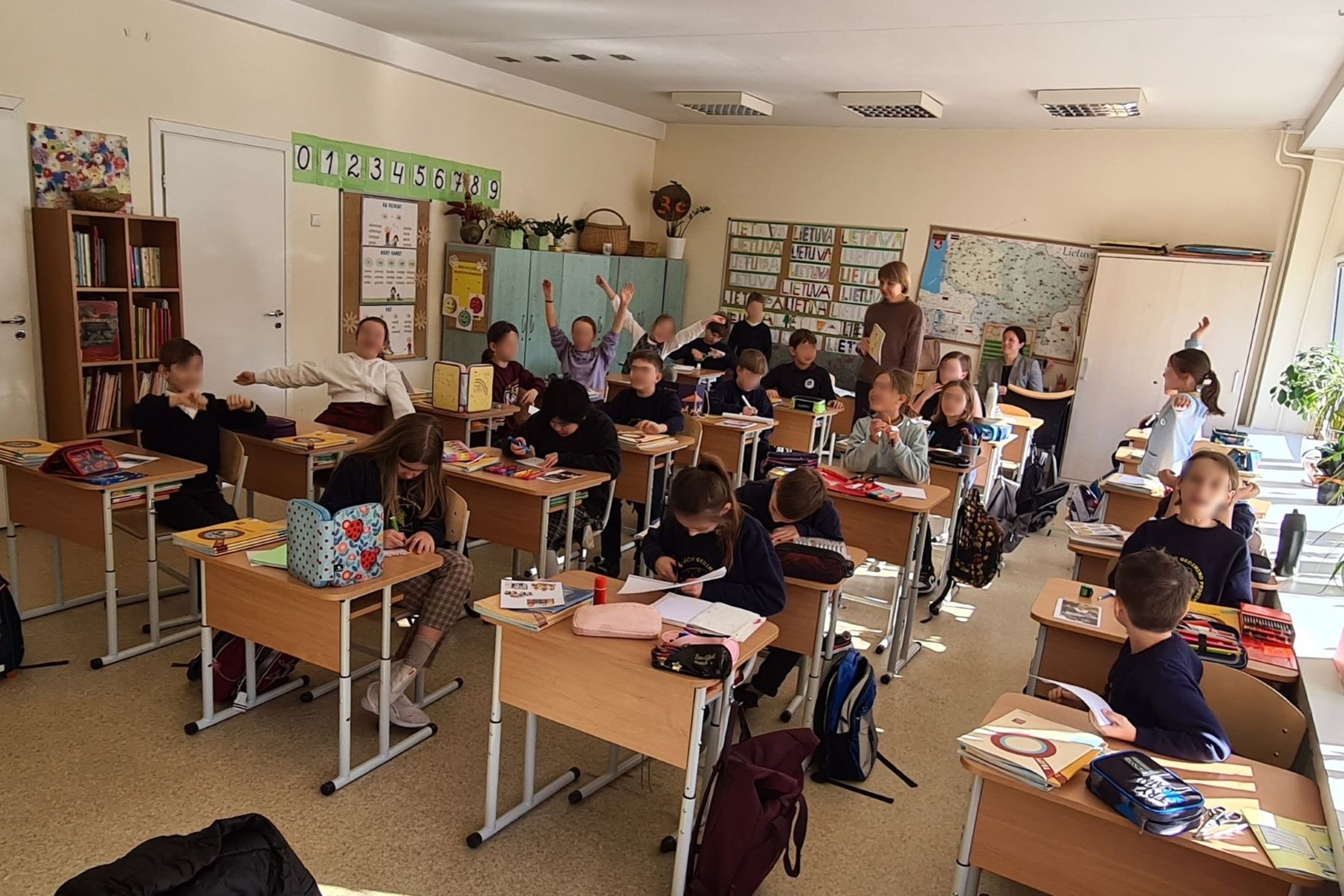 Mokiniai sėdi klasės suoluose ir mojuoja rankomis.