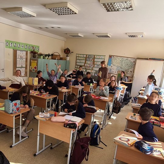 Mokiniai sėdi klasės suoluose ir mojuoja rankomis.