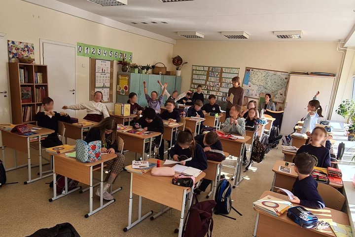 Mokiniai sėdi klasės suoluose ir mojuoja rankomis.
