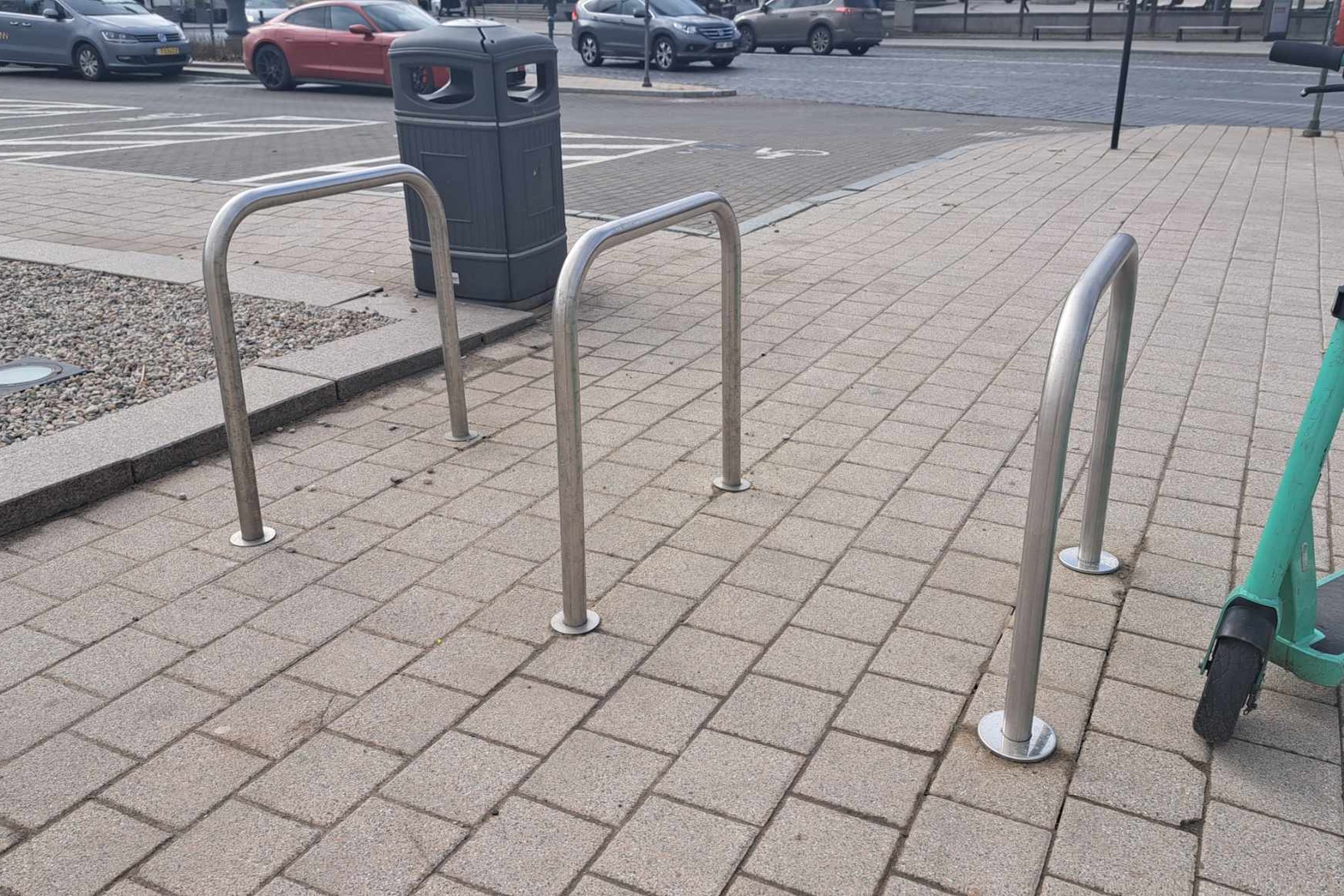 Nufotografuoti dviračių stulpai jau ištiesinti. Fone matosi šiukšliadėžė ir stovėjimo aikštelė.