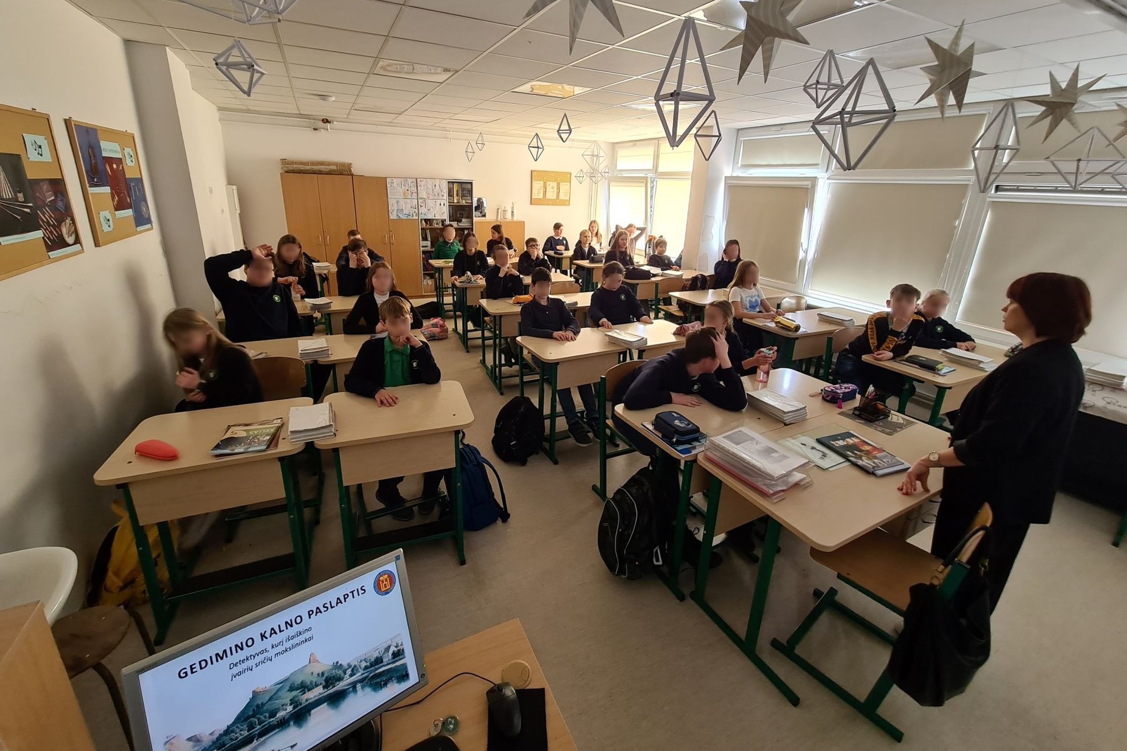 Nuotrauka, kurioje matosi penktokų klasė, prieš juos stovinti mokytoja ir klasės kompiuterio ekrane žibantis užrašas Gedimino kalno paslaptis.