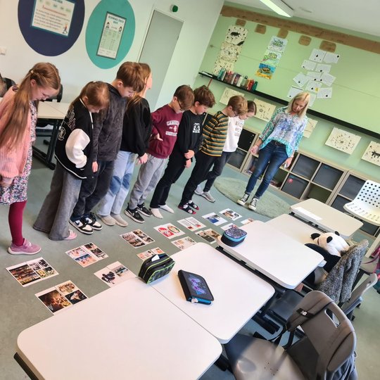 Mokiniai, sustoję klasėje, žiūri į ant žemės padėtas fotografijas, spausdintas baltuose lapuose.