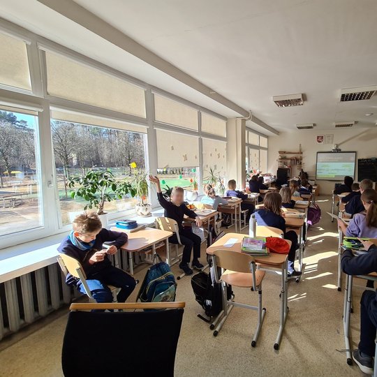 Mokiniai sėdi klasės suoluose ir mojuoja rankomis.