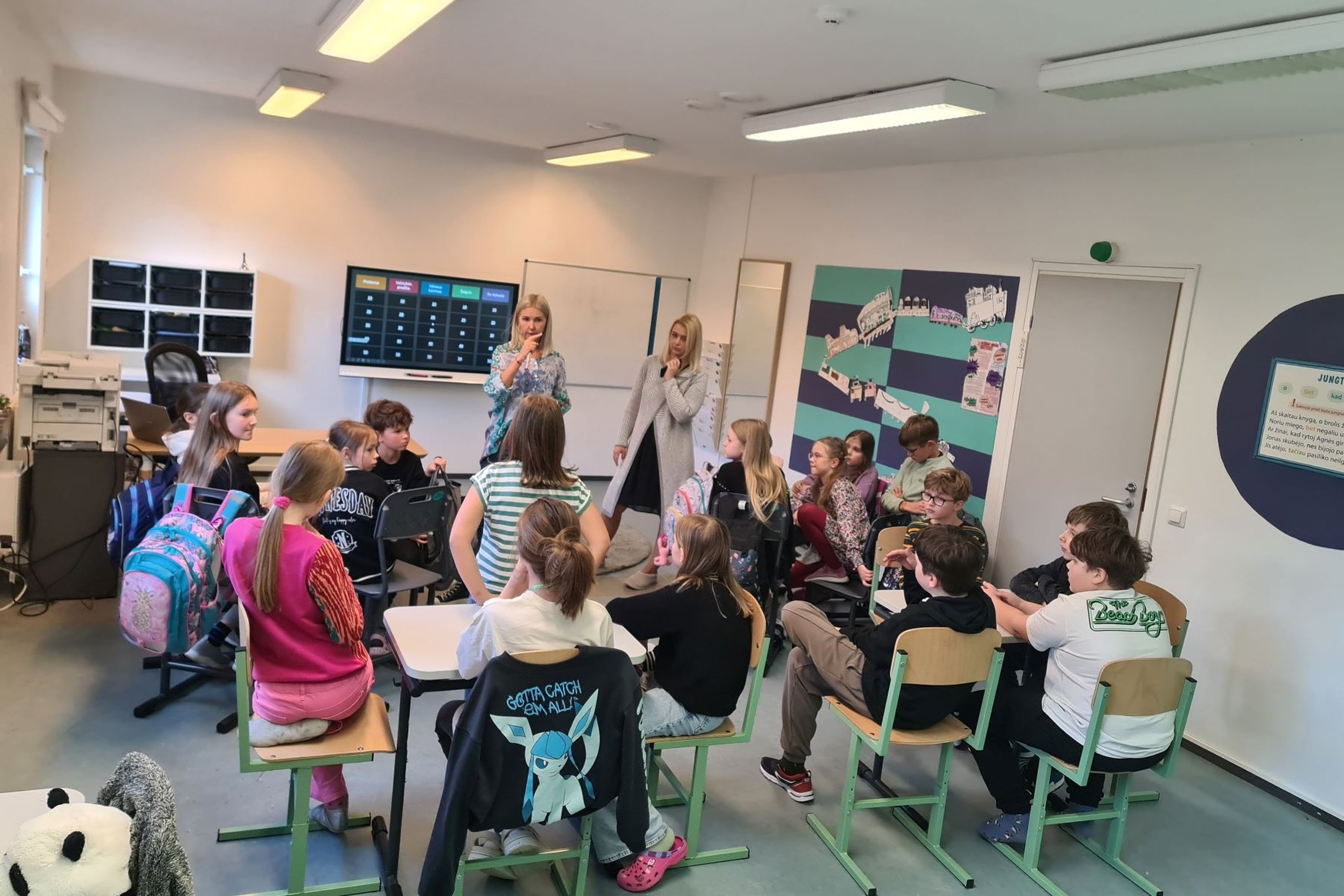Klasėje mokiniai, susėdę ant klasės kėdžių. Prieš juos dvi mokytojos.