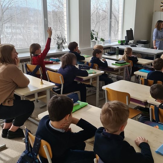 Kol kiti vaikai sėdi suoluose, viena mergaitė kelia ranką, nori užduoti klausimą edukatorei.