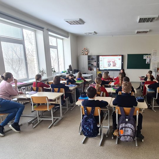 Fotografuota edukacinė pamoka iš klasės galo. Visi sėdi suoluose, net mokytoja.