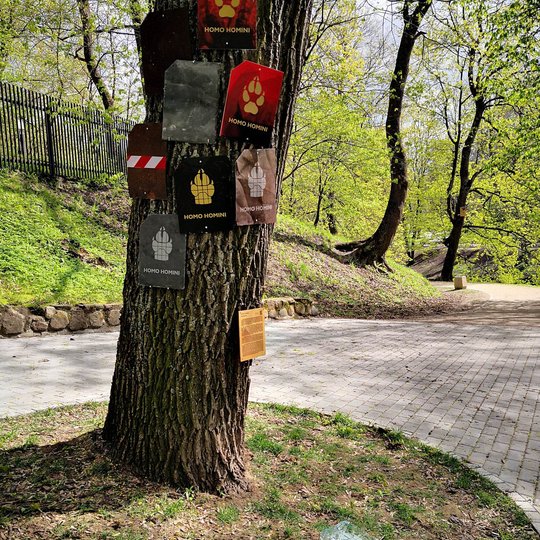 "Homo Homini" instaliacija kurta iš akmeninių ir medinių plokštelių, sukabintų prie medžio. Vaizduoja vilko pėdas.