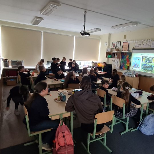 Vaikai klasėje. Vieni žiūri į projektorių, kiti į objektyvą, visi juda, dirba, mąsto...