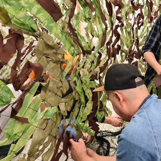 Du direkcijos darbuotojai riša maskuojantį tinklą. Viena iš darbuotojų juostas persimetusi per petį.