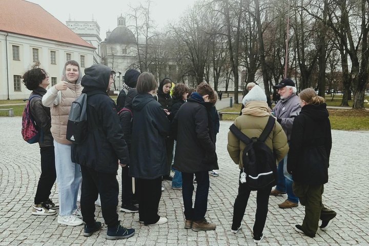 Nuotraukoje mokiniai įdėmiai klausosi edukatoriaus Stanislovo pasakojimo apie Karaliaus Mindaugo paminklą ir karaliaus istoriją.