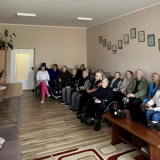 Visa grupelė globos namų gyventojų sėdi svetainėje ir klausosi edukacijos apie Gediminą, Gediminaičius ir Vilniaus pilis.