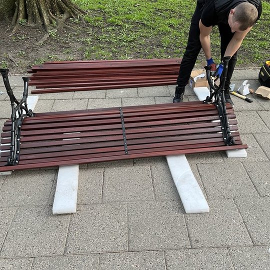 Vienas iš meistrų suka varžtus į lauko suolo atramą. Suolas paguldytas ant putplasčio.