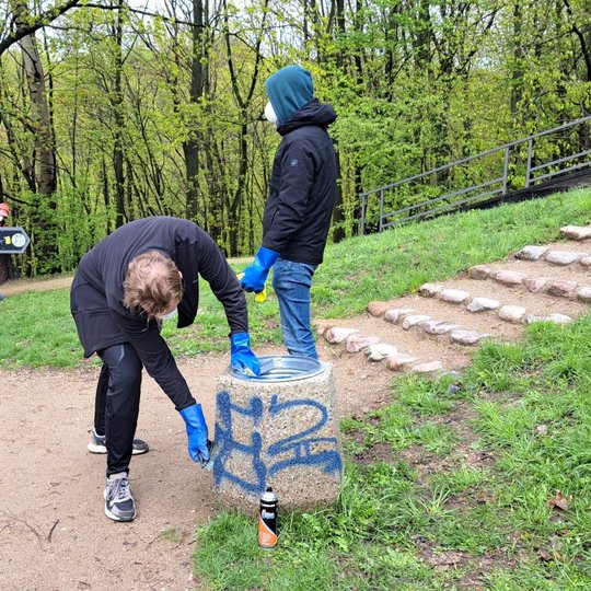 Vienas savanoris, pasilenkęs prie šiukšliadėžės, šepečiu ir chemija bando nuvalyti graffiti piešinį.