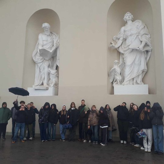 Aštuntokų grupė išsirikiavę su skėčiais prie Arkikatedros fasado. Virš jų matosi statulos.