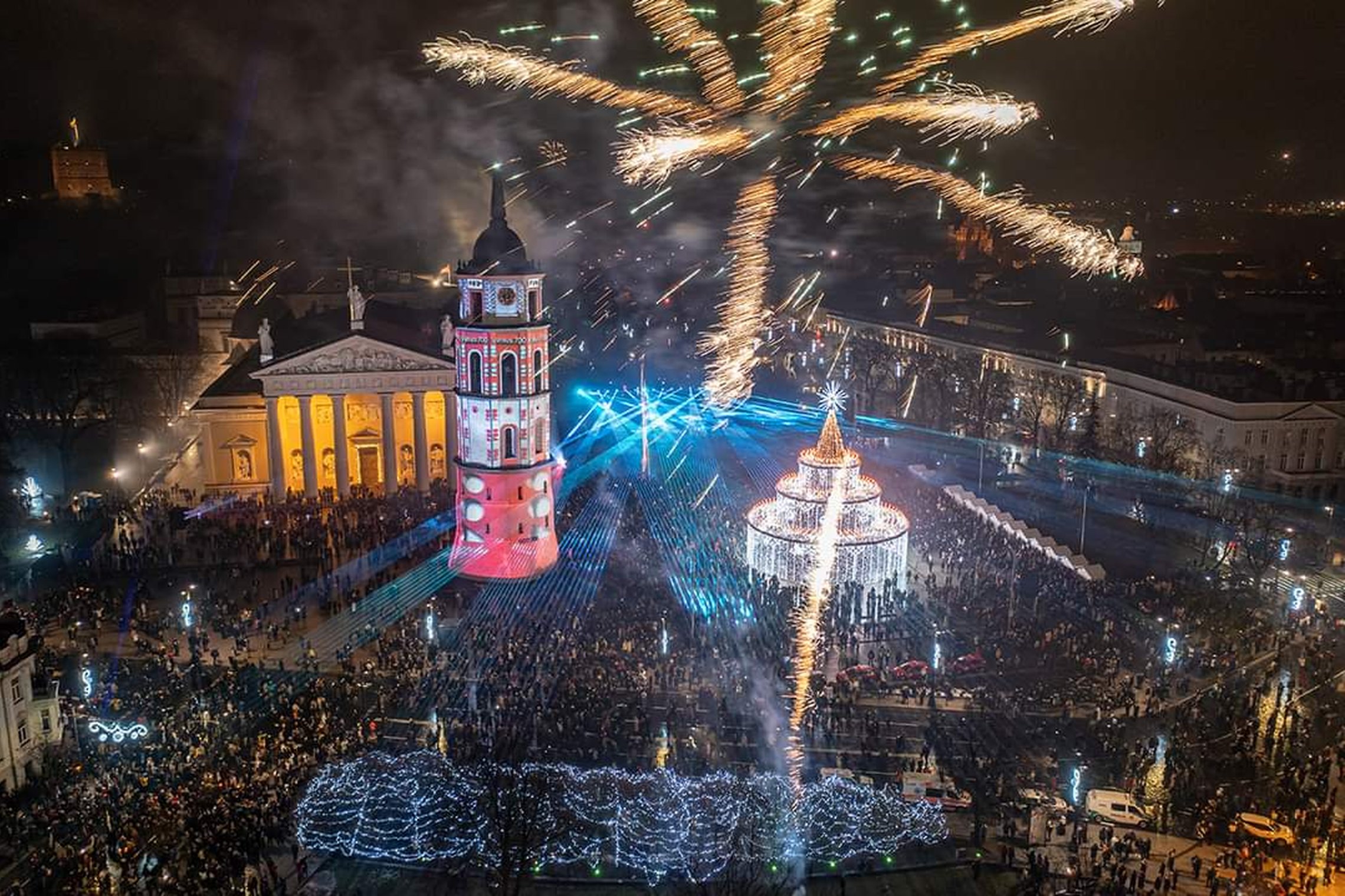 Naujametinė Žemutinės pilies nuotrauka iš viršaus. Matosi kalėdinė eglutė katedros aikštėje, apšviestas varpinės bokštas, taip pat Vilniaus katedra. Nuotrauka kupina žmonių bei fejerverkų.