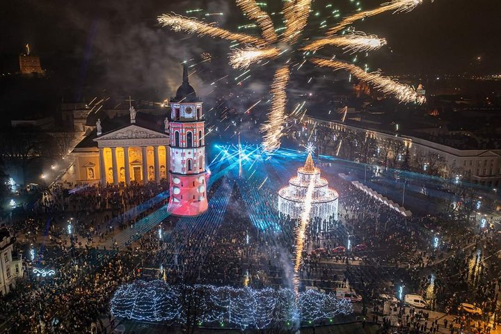 Naujametinė Žemutinės pilies nuotrauka iš viršaus. Matosi kalėdinė eglutė katedros aikštėje, apšviestas varpinės bokštas, taip pat Vilniaus katedra. Nuotrauka kupina žmonių bei fejerverkų.