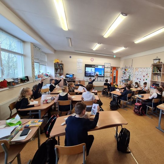 Nufotografuota klasė. Mokiniai matosi iš nugarų. Jie atlieka pavestas užduotis.