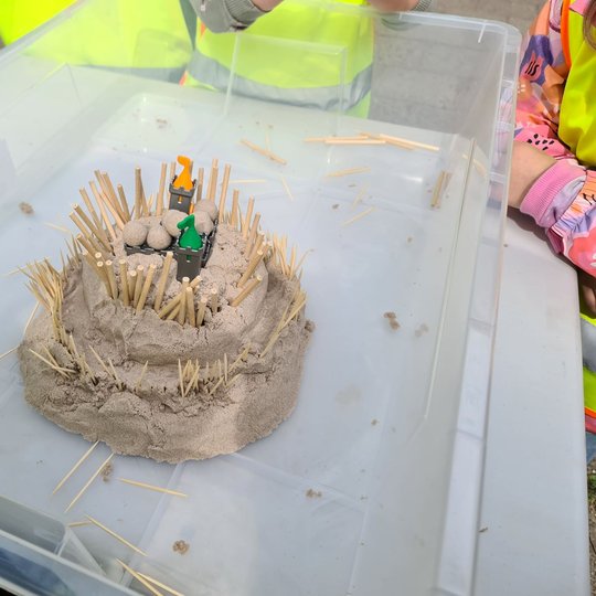 Vaikų pagaminta smėlio pilis su mediniais pagaliukais plastikinėje dėžėje.