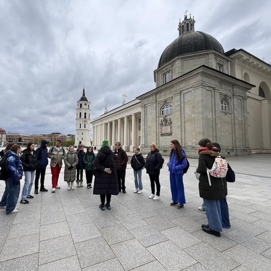 Jaunieji turistai išsirikiavę puslankiu aplink gidę Katedros aikštėje.