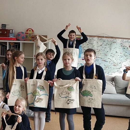 Vilniaus Maironio progimnazijos pradinukai stovi klasėje ir pozuoja su dovanų gautais medžiaginiais maišeliais.