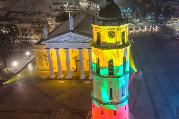 Katedra, jos varpinė ir Gedimino kalnas su pilimi nufotografuota iš oro, vakarą. Aplink daug šviesų, o katedros varpinė apšviesta trispalvės spalvomis.