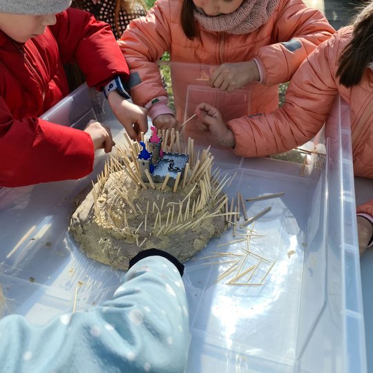 Plastikinėje dėžėje smėlio pilis, susmaigstyta pieštukais. Ją liečia keli mokiniai.