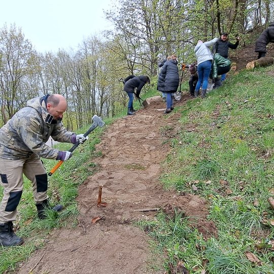 Pirmame plane vyras kala kuolą prie skersinio, antrame plane jo kolegos tvirtina skersiniu aukščiau tako.