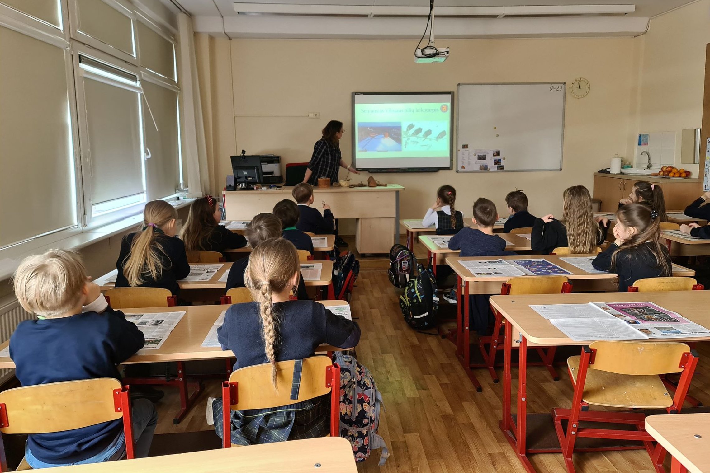 Nufotografuota klasė. Nuotrauka daryta iš klasės galo. Pamokoje sėdi mokiniai, klausosi edukatorės vedamos paskaitos.