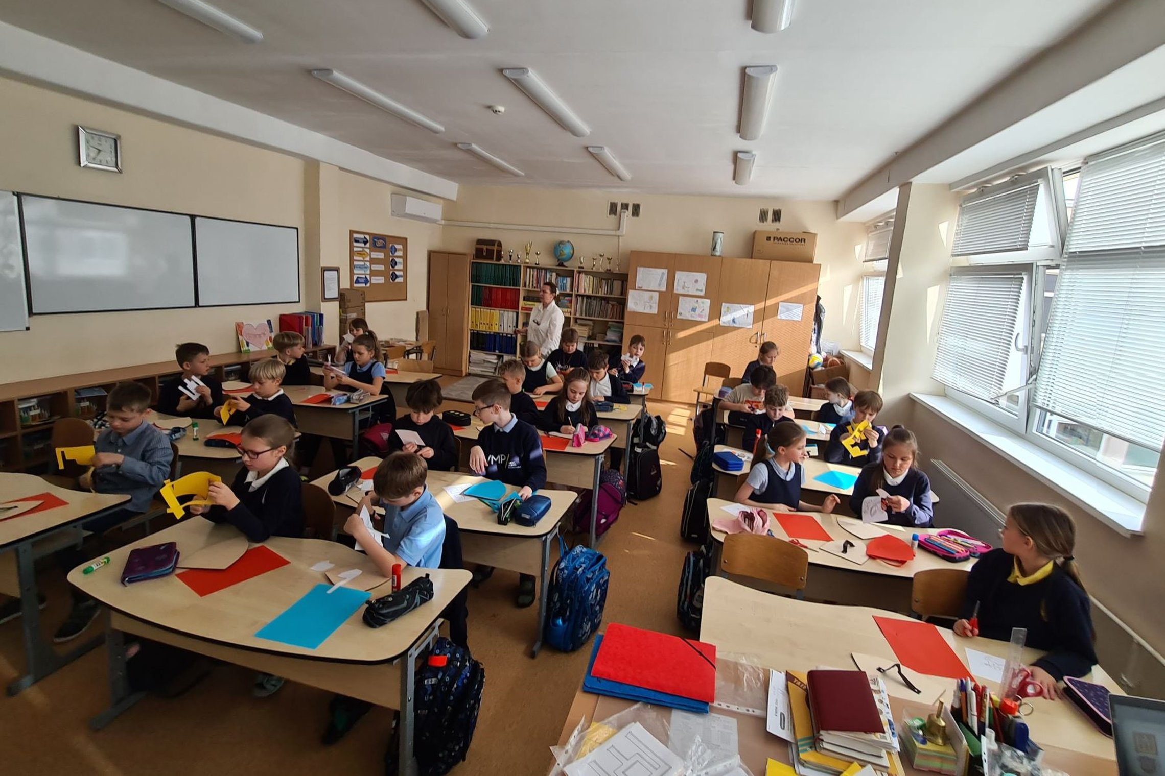 Nufotografuota mokinių klasė. Mokiniai suoluose karpo ir klijuoja spalvotą popierių.