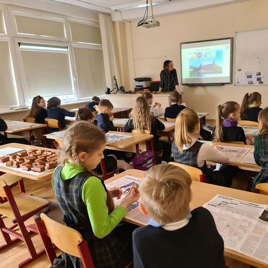 Nufotografuota klasė mokinių, besiklausančių paskaitos. Klasės gale išdėlioti molio gabaliukai - vaikai lipdys puodus.