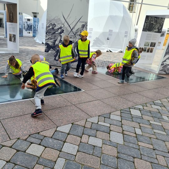 Vaikai stebi Valdovų rūmų rūsį per kieme esantį stiklą.
