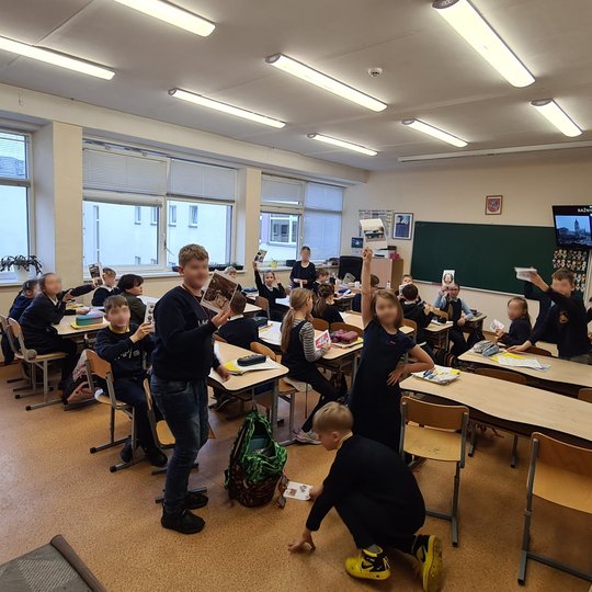 Nufotografuota klasė, pilna mokinių. Fotografuota iš klasės galo. Vaikai iškėlę rankas, rankose laiko piešinius.