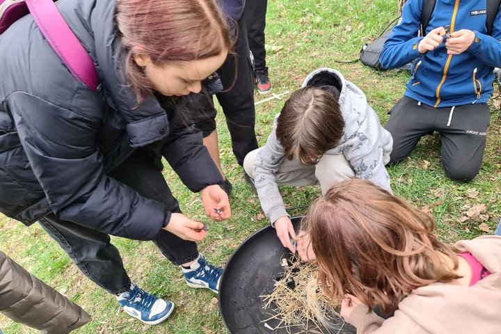 Trys mokiniai bando užkurti ugnį paruoštoje lėkštėje su šiaudais, besinaudodami skiltuvais.