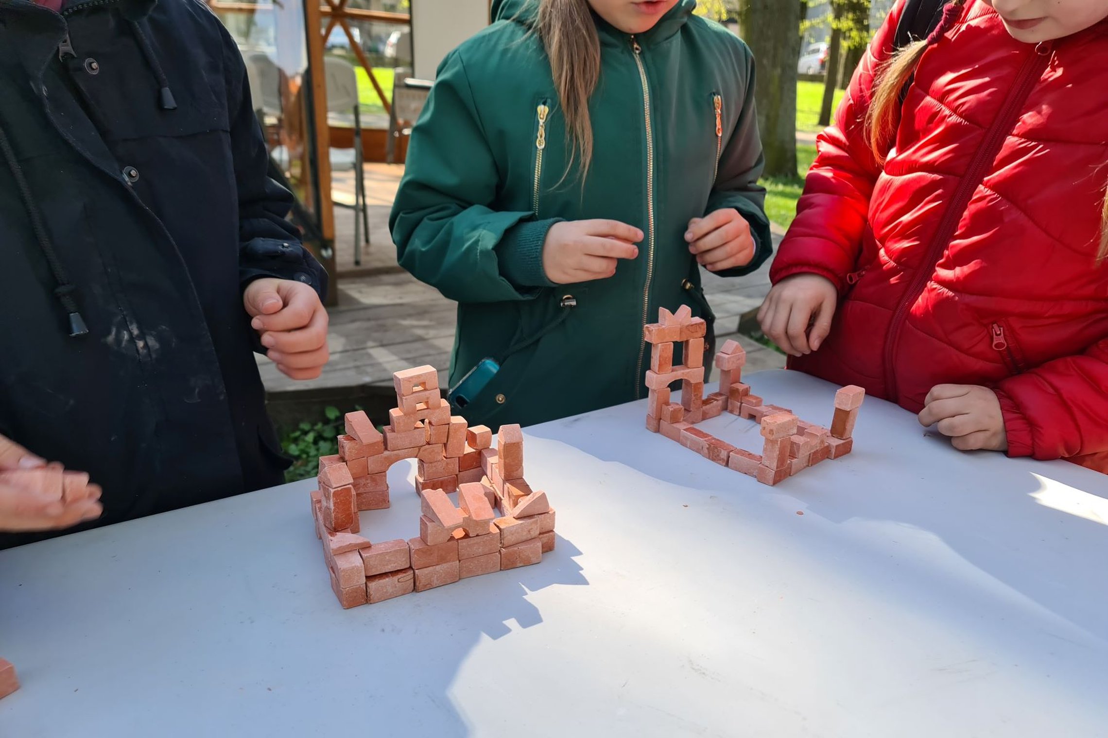 Ant stalo stovi dvi iš mažų plytelių sudėliotos pilys. Šalia jų - trijų mokinių siluetai.