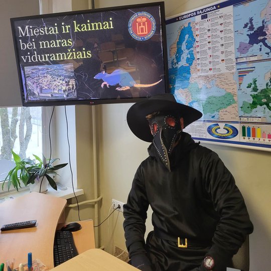 Edukatorius su maro daktaro kostiumu sėdi prie stalo. Virš jo televizoriaus ekranas, kuriame pristatomas maras.