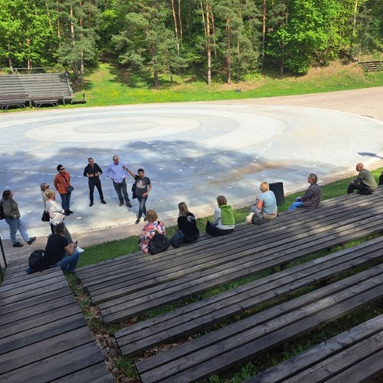Klausoma ekskursija Kalnų parko estradoje. Žmonės sėdi ant suolų.