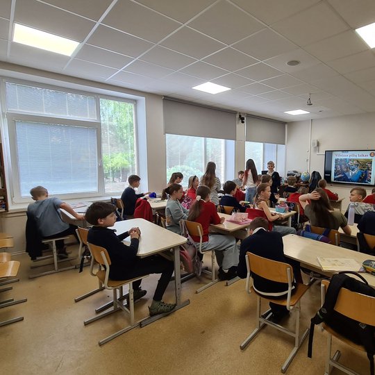 Klasėje šurmuliuoja vaikai, priekyje matosi per projektorių rodomos skaidrės.