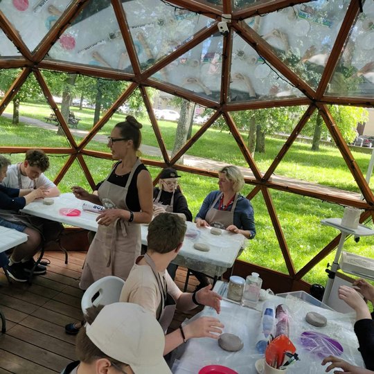 Žmonės lipdo molį, o edukatorė jiems pasakoja instrukcijas.