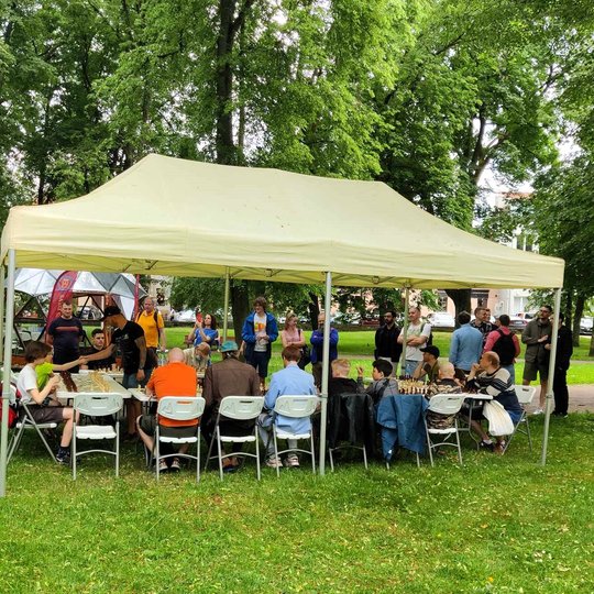 Po paviljonu prie stalų sėdi šachmatininkai. Minia žmonių stovi aplink juos.